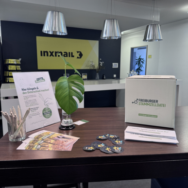 Theke mit Inxmail-Logo, Karton 'Freiburger Stammzelldatei', Flyer, Abzeichen und Monstera-Blatt