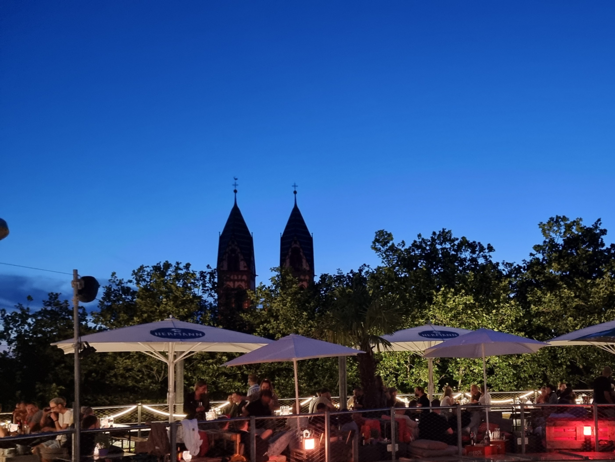 Abendliche Terrasse mit Gästen unter Sonnenschirmen vor zwei Kirchtürmen