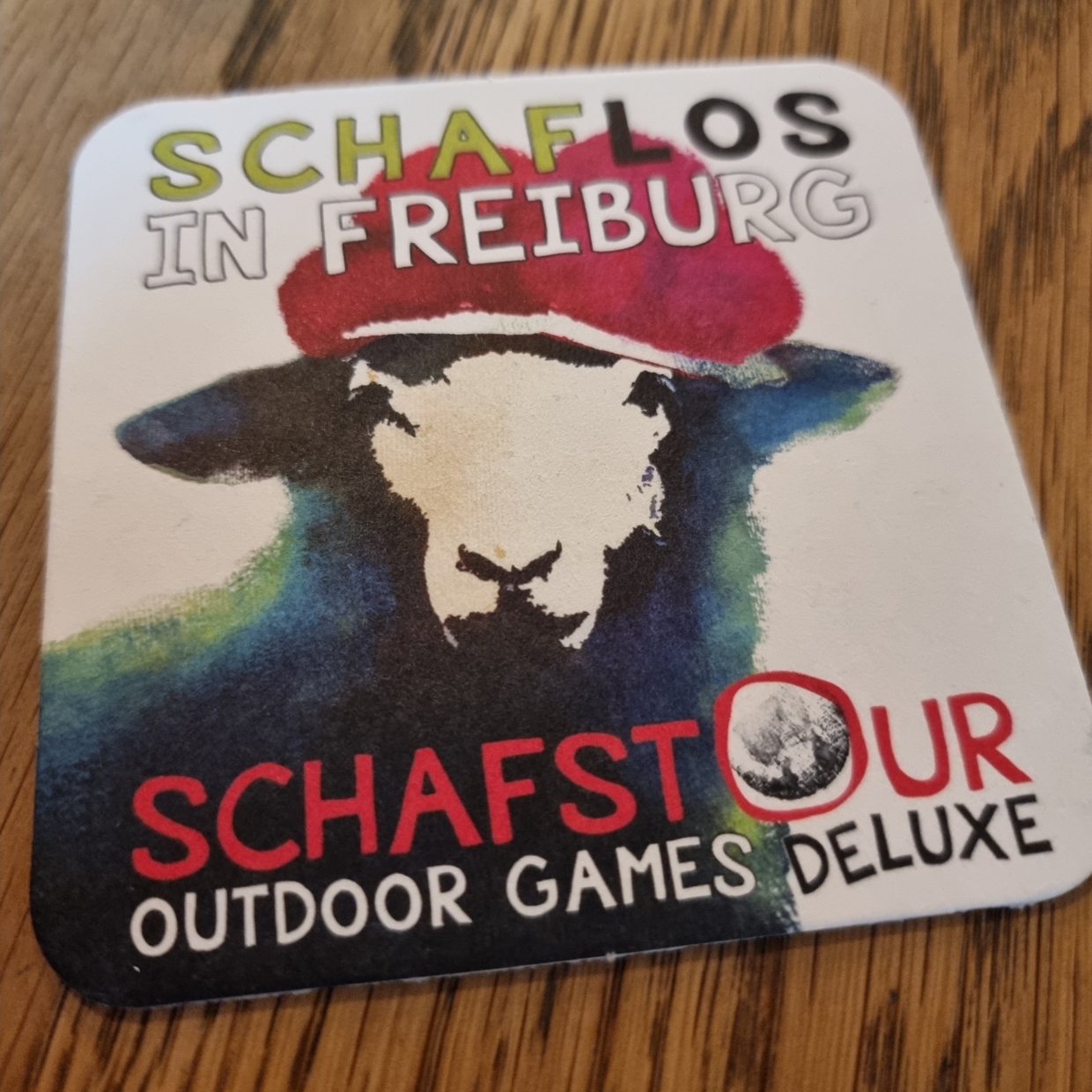 Untersetzer mit Schaf-Illustration, Text 'SCHAFLOS IN FREIBURG' und 'SCHAFSTOUR OUTDOOR GAMES DELUXE' auf Holz.